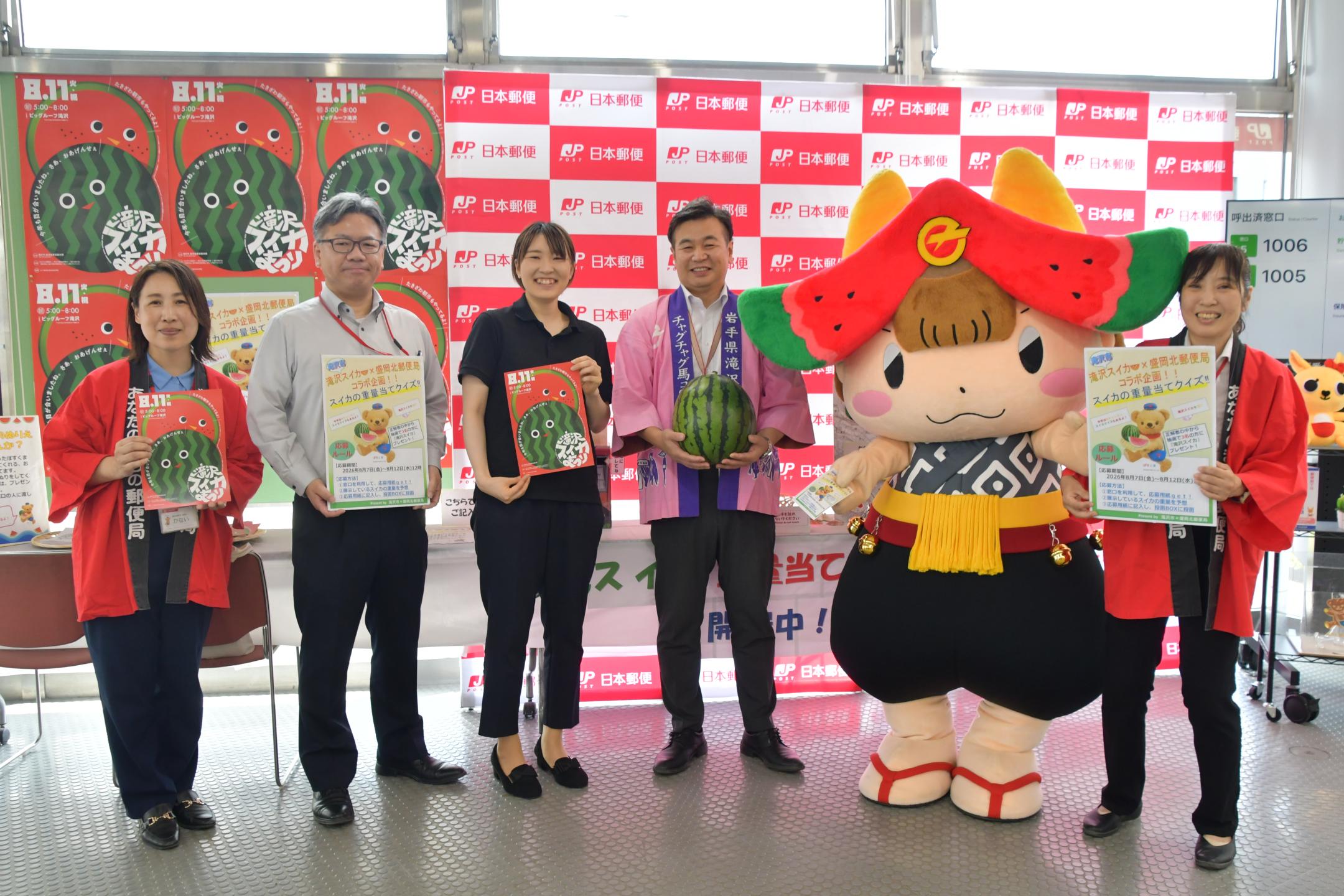 スイカまつりポスター、スイカ重量当てクイズポスター、滝沢スイカなどを手にする局員と市農林課職員、ちゃぐぽん